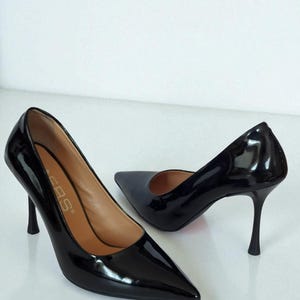 May include: A pair of glossy black high-heeled shoes with pointed toes. The heels are slender and approximately 7.6 cm tall. The shoes have a sleek, elegant design, suitable for formal occasions.