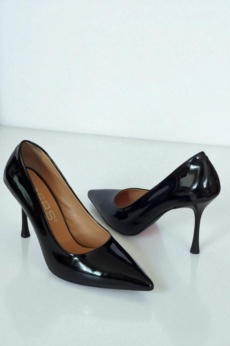 May include: A pair of glossy black high-heeled shoes with pointed toes. The heels are slender and approximately 7.6 cm tall. The shoes have a sleek, elegant design, suitable for formal occasions.