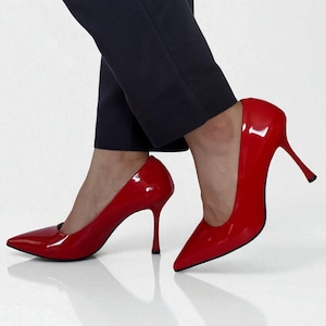 May include: A pair of glossy red high-heeled shoes with pointed toes. The stiletto heels are slender and the shoes have a patent leather finish. The shoes are paired with dark navy blue trousers.