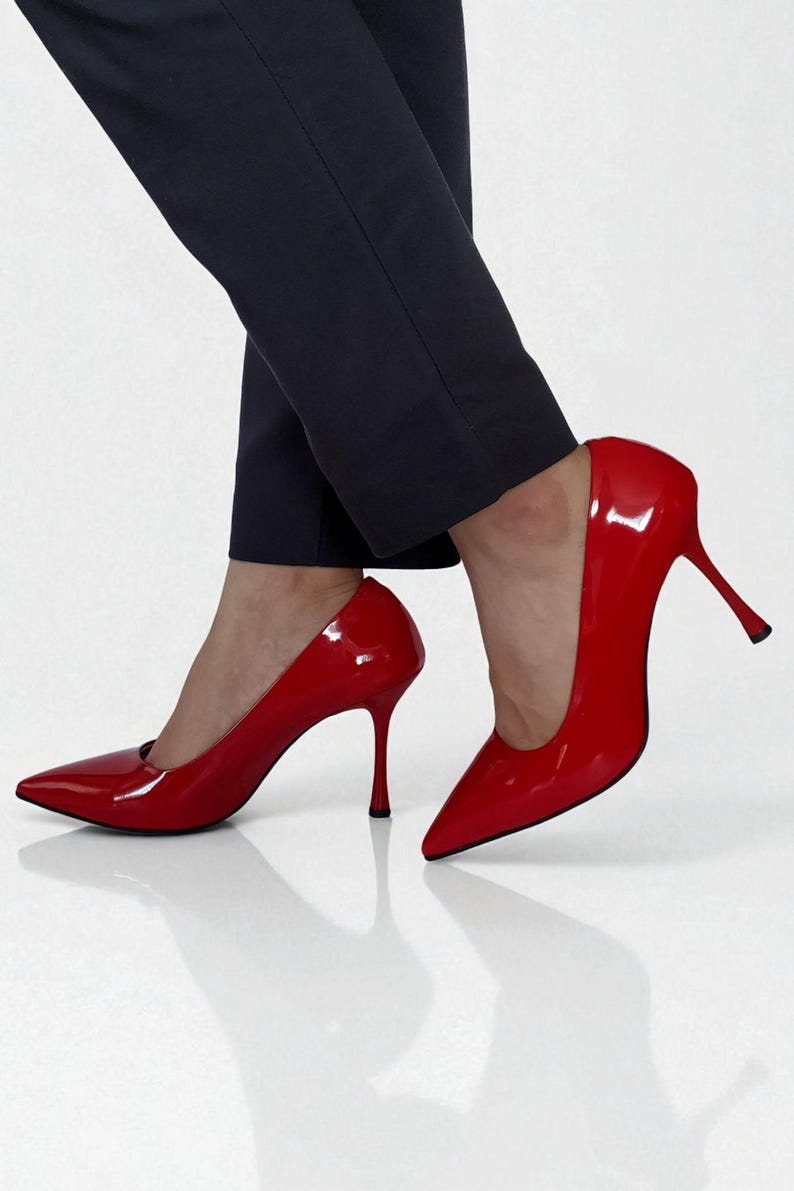 May include: A pair of glossy red high-heeled shoes with pointed toes. The stiletto heels are slender and the shoes have a patent leather finish. The shoes are paired with dark navy blue trousers.