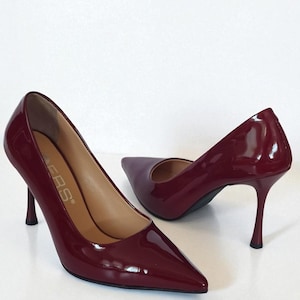 May include: A pair of burgundy patent leather high heels with pointed toes and stiletto heels. The shoes have a glossy finish and are set against a white background. The heels are approximately 10 cm tall.