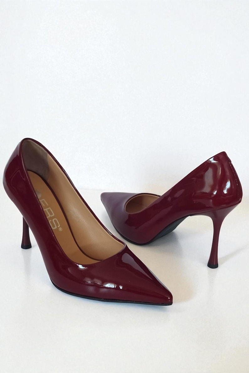 May include: A pair of burgundy patent leather high heels with pointed toes and stiletto heels. The shoes have a glossy finish and are set against a white background. The heels are approximately 10 cm tall.