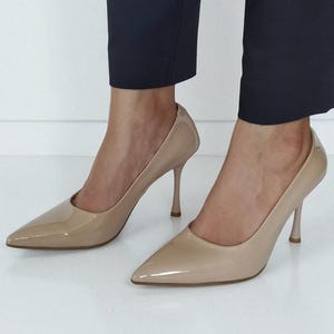 May include: Beige high-heeled pumps with a pointed toe and a glossy finish. The shoes feature a slender stiletto heel, approximately 8.9 cm high. The shoes are worn with navy blue trousers.