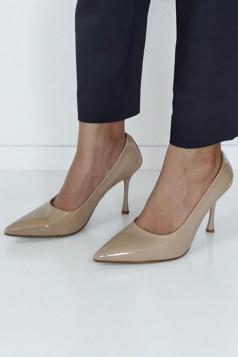 May include: Beige high-heeled pumps with a pointed toe and a glossy finish. The shoes feature a slender stiletto heel, approximately 8.9 cm high. The shoes are worn with navy blue trousers.