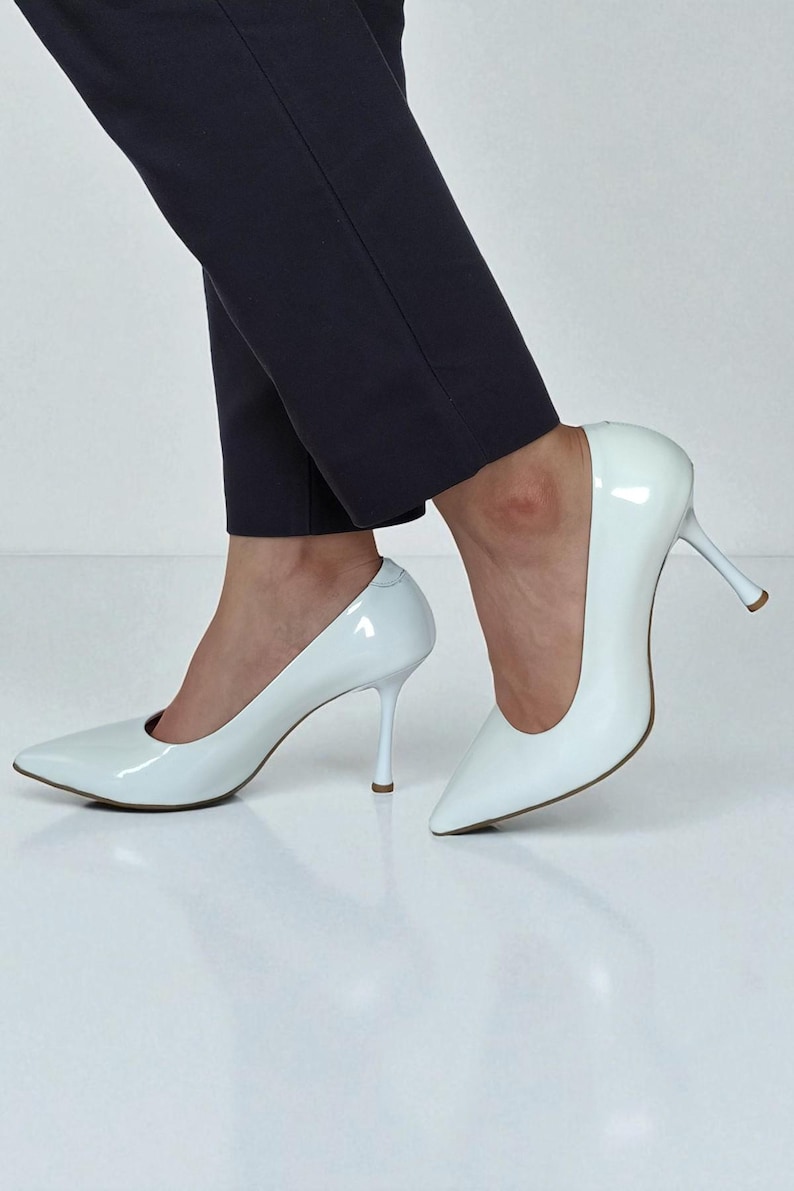 May include: White patent leather pointed-toe court shoes with stiletto heels. The shoes have a glossy finish and a classic design. The heels are approximately 7.6 cm tall. The shoes are paired with dark navy blue trousers.