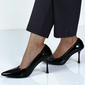 May include: A pair of glossy black stiletto heels with pointed toes. The shoes have a high heel and are worn with dark navy blue cropped trousers. The shoes reflect the bright white surface they are on.