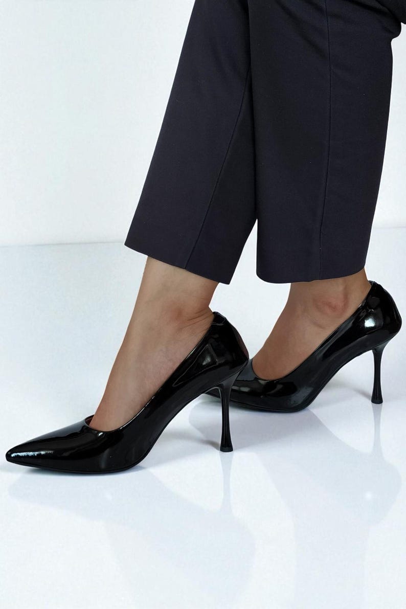 May include: A pair of glossy black stiletto heels with pointed toes. The shoes have a high heel and are worn with dark navy blue cropped trousers. The shoes reflect the bright white surface they are on.