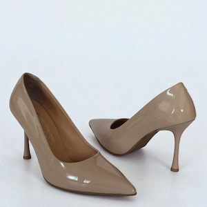 May include: A pair of beige patent leather high heels with pointed toes and stiletto heels. The shoes are set against a white background, showcasing their glossy finish and elegant design. The heels are approximately 7.6 cm tall.