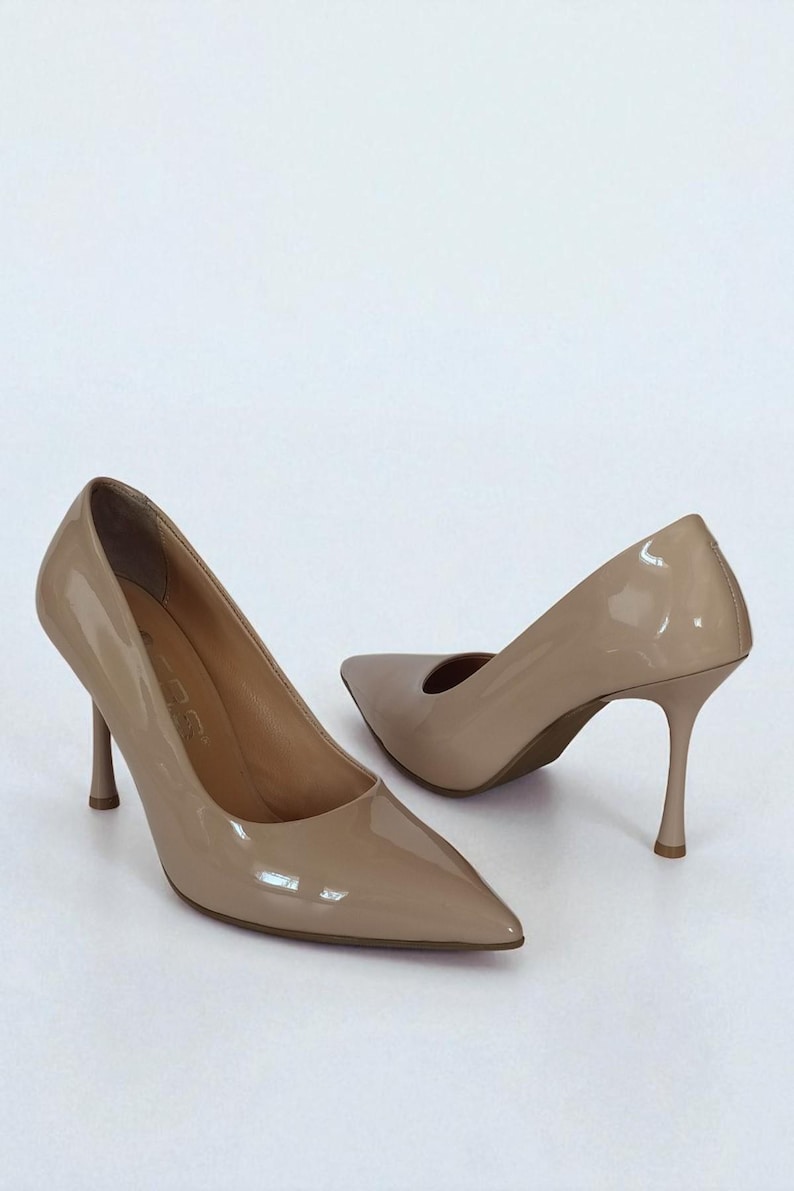 May include: A pair of beige patent leather high heels with pointed toes and stiletto heels. The shoes are set against a white background, showcasing their glossy finish and elegant design. The heels are approximately 7.6 cm tall.
