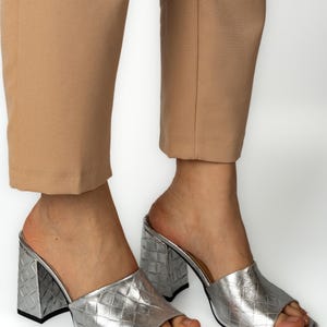 May include: Silver metallic open-toe mules with a textured, woven-like pattern. The shoes have a block heel and a square toe. The image shows the shoes on a person wearing tan pants.