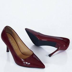 May include: Burgundy patent leather court shoes with pointed toes and stiletto heels. The shoes have a glossy finish and a classic design, suitable for formal occasions. The heels are approximately 7.6 cm high.