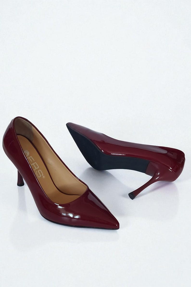 May include: Burgundy patent leather court shoes with pointed toes and stiletto heels. The shoes have a glossy finish and a classic design, suitable for formal occasions. The heels are approximately 7.6 cm high.