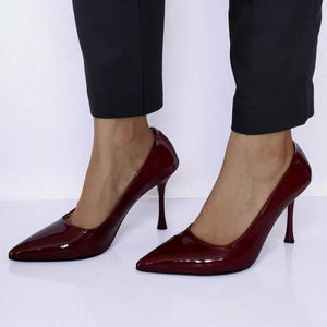 May include: Burgundy patent leather high heels with pointed toes and stiletto heels. The shoes are worn with dark trousers. The heels have a sleek, elegant design, suitable for formal occasions.