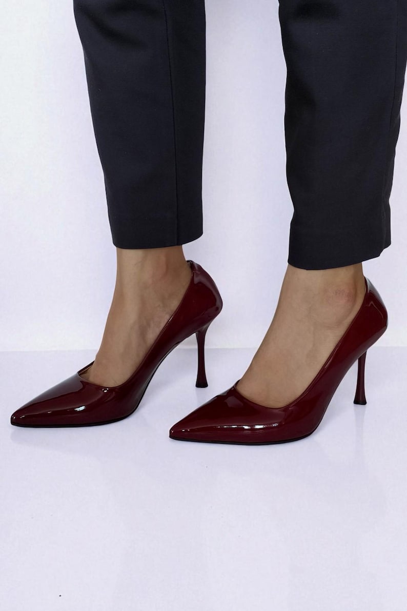 May include: Burgundy patent leather high heels with pointed toes and stiletto heels. The shoes are worn with dark trousers. The heels have a sleek, elegant design, suitable for formal occasions.