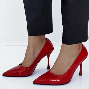 May include: A pair of glossy red high-heeled pumps with pointed toes. The shoes have a stiletto heel and are worn with black trousers. The shoes are a vibrant shade of red.