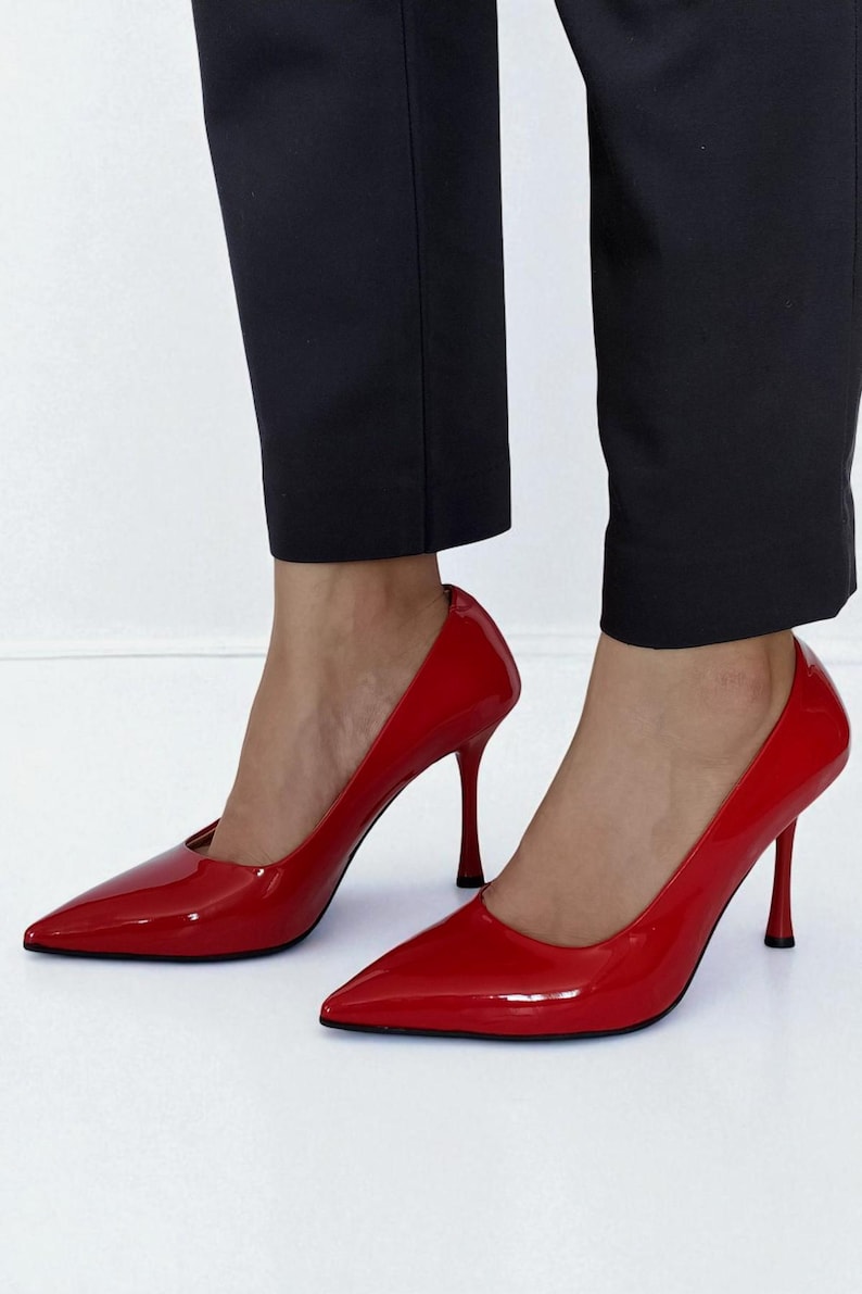 May include: A pair of glossy red high-heeled pumps with pointed toes. The shoes have a stiletto heel and are worn with black trousers. The shoes are a vibrant shade of red.