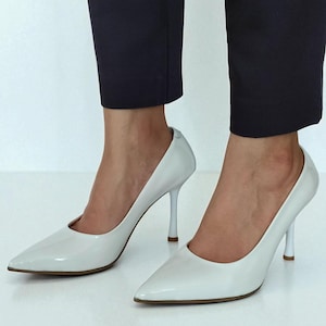 May include: A pair of white, pointed-toe high heels. The shoes have a glossy finish and slender heels. The heels are approximately 10 cm tall. The shoes are worn with dark navy blue trousers.