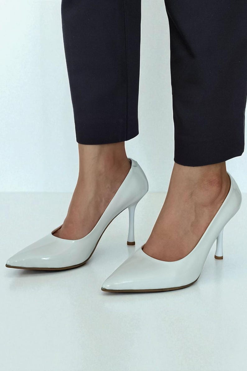 May include: A pair of white, pointed-toe high heels. The shoes have a glossy finish and slender heels. The heels are approximately 10 cm tall. The shoes are worn with dark navy blue trousers.