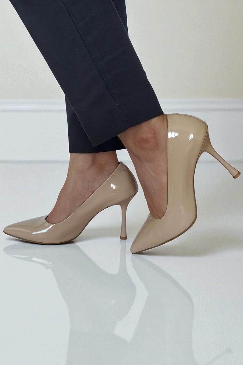 May include: Beige patent leather high heels with pointed toes. The stiletto heels are slender and elegant. The shoes are worn with dark navy blue trousers. The glossy finish reflects light.
