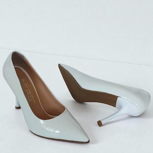 May include: A pair of pale blue high-heeled shoes with pointed toes. The heels are thin and the soles are brown. The shoes have a glossy finish and are set against a white background. The brand name is visible on the insole.