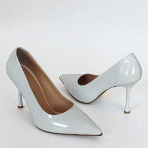 May include: A pair of white high-heeled pumps with pointed toes. The shoes have a glossy finish and slender heels. The interior of the shoes is a light tan colour. The heels are approximately 7.6 cm high.