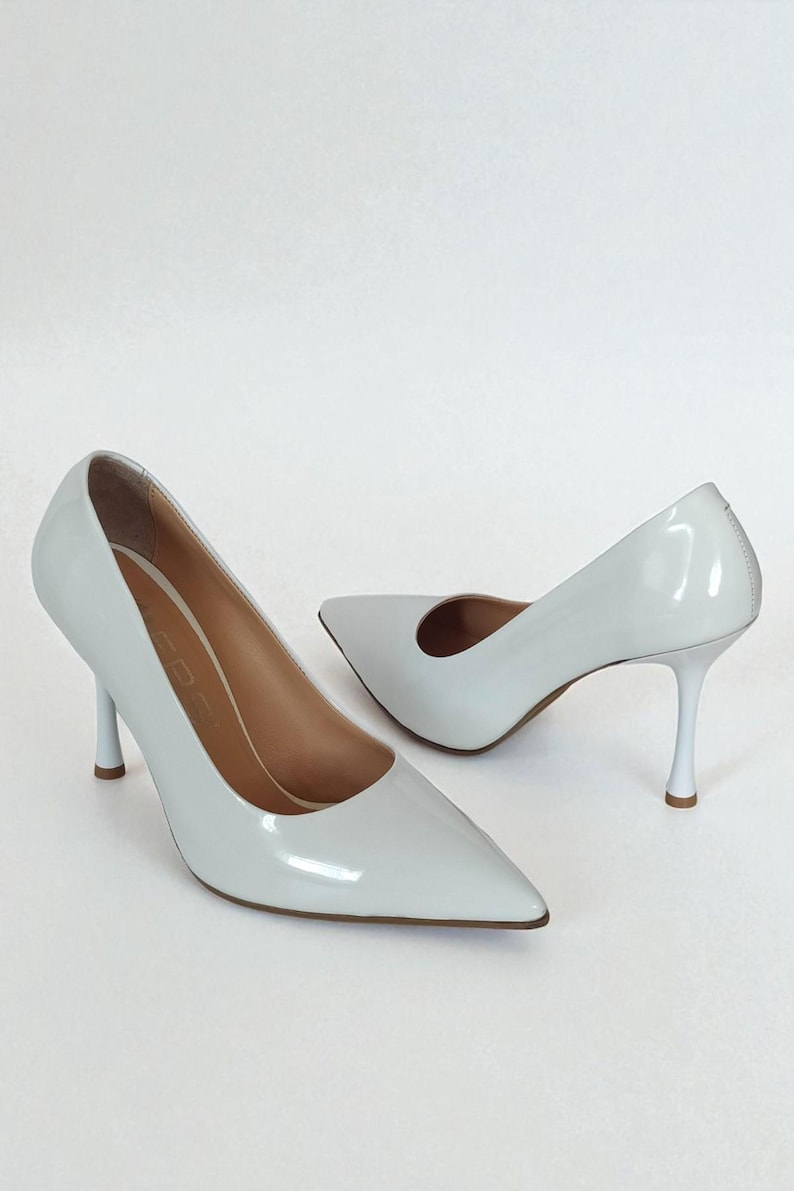 May include: A pair of white high-heeled pumps with pointed toes. The shoes have a glossy finish and slender heels. The interior of the shoes is a light tan colour. The heels are approximately 7.6 cm high.