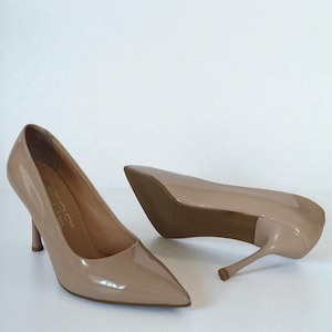 May include: A pair of beige high-heeled shoes with pointed toes. The shoes have a glossy finish and slender heels. The heels appear to be approximately 7.6 cm tall. The shoes are set against a white background.