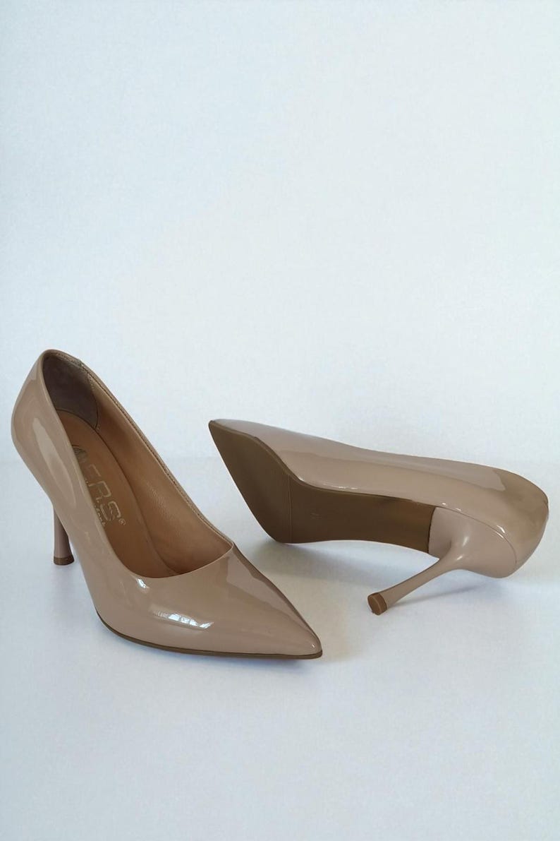 May include: A pair of beige high-heeled shoes with pointed toes. The shoes have a glossy finish and slender heels. The heels appear to be approximately 7.6 cm tall. The shoes are set against a white background.