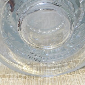 May include: Clear glass object with a textured surface and a circular design. The text "Made in Portugal" is visible. The object is sitting on a beige woven surface.