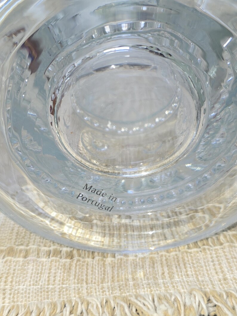 May include: Clear glass object with a textured surface and a circular design. The text "Made in Portugal" is visible. The object is sitting on a beige woven surface.