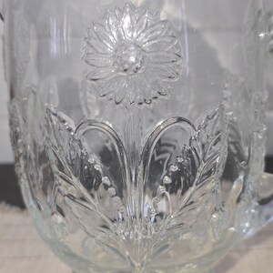May include: Clear glass pitcher with embossed floral and leaf design. The pitcher features a large sunflower in the center and detailed leaf patterns around the base. It has a handle and a spout for pouring.