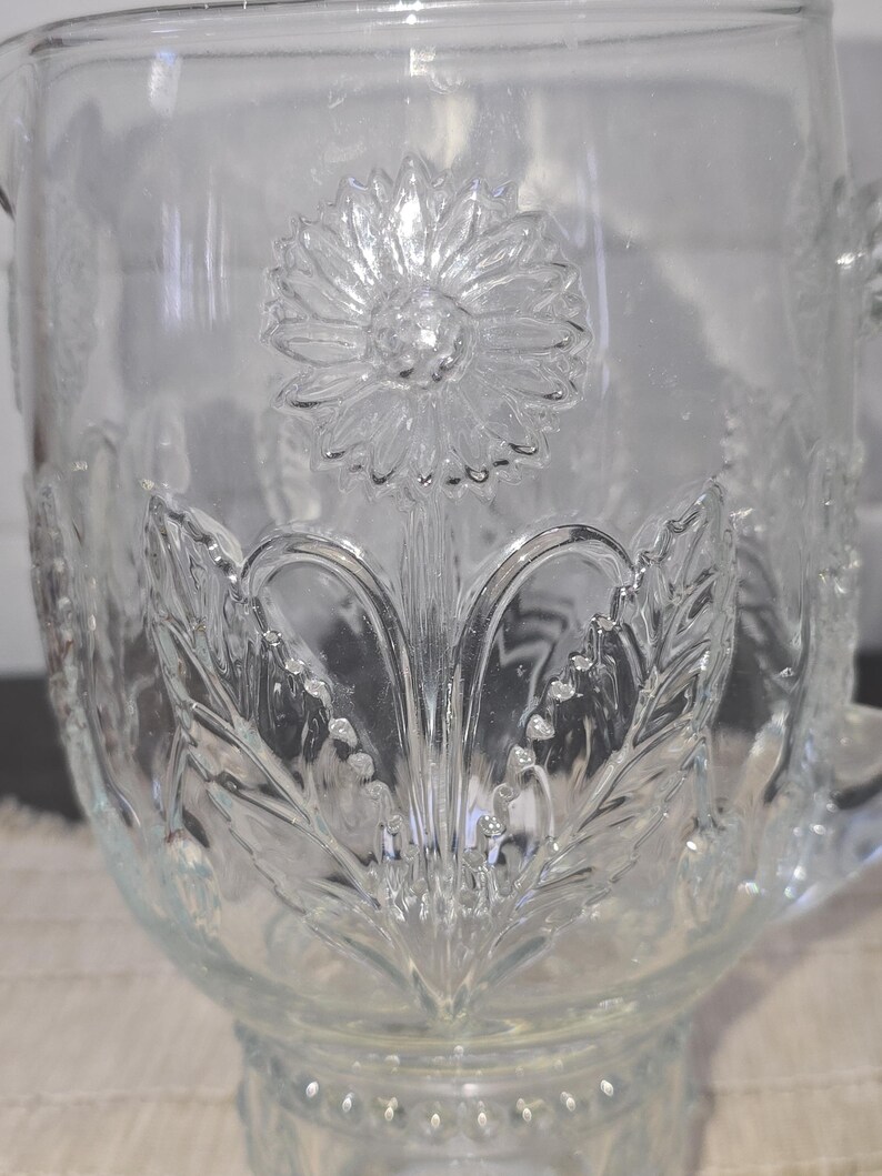 May include: Clear glass pitcher with embossed floral and leaf design. The pitcher features a large sunflower in the center and detailed leaf patterns around the base. It has a handle and a spout for pouring.