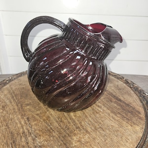 May include: A dark purple glass pitcher with a swirl design and a handle. The pitcher is sitting on a wooden surface.