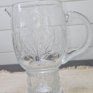 May include: Clear glass pitcher with a floral design. The pitcher has a spout and a handle. The floral design is embossed on the glass. The pitcher sits on a decorative base.