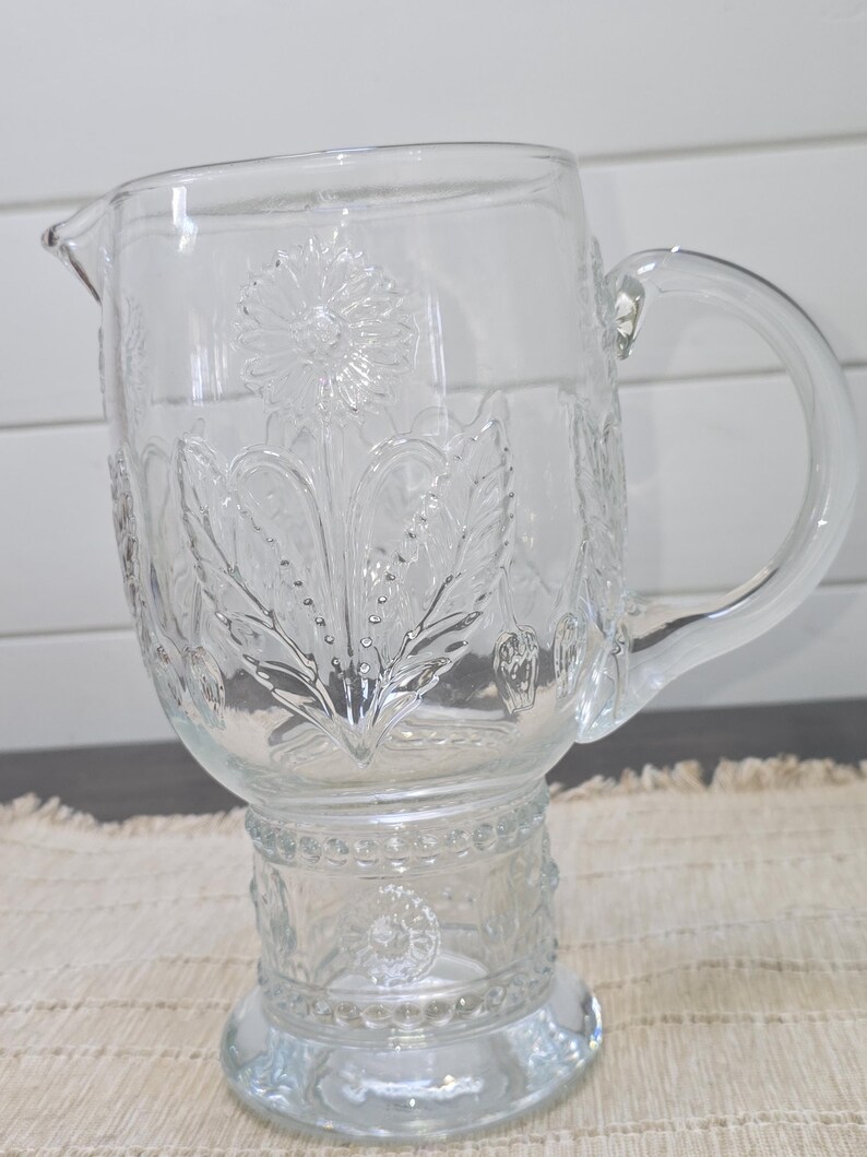 May include: Clear glass pitcher with a floral design. The pitcher has a spout and a handle. The floral design is embossed on the glass. The pitcher sits on a decorative base.