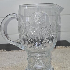 May include: Clear glass pitcher with a handle and a spout. The pitcher features an embossed floral design, including a large flower in the center and surrounding leaves. The pitcher sits on a decorative base.