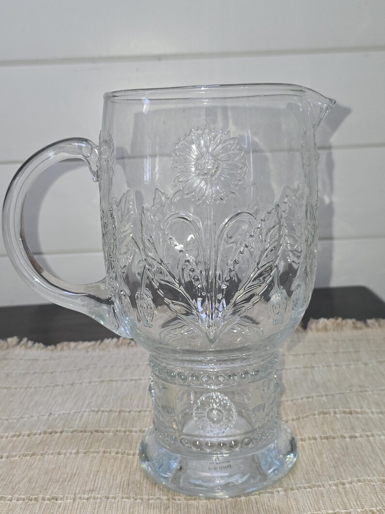May include: Clear glass pitcher with a handle and a spout. The pitcher features an embossed floral design, including a large flower in the center and surrounding leaves. The pitcher sits on a decorative base.