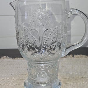 May include: Clear glass pitcher with a floral design. The pitcher features a spout and a handle. The floral design is embossed on the glass, with a large flower in the center and leaves around it. The pitcher sits on a pedestal base.