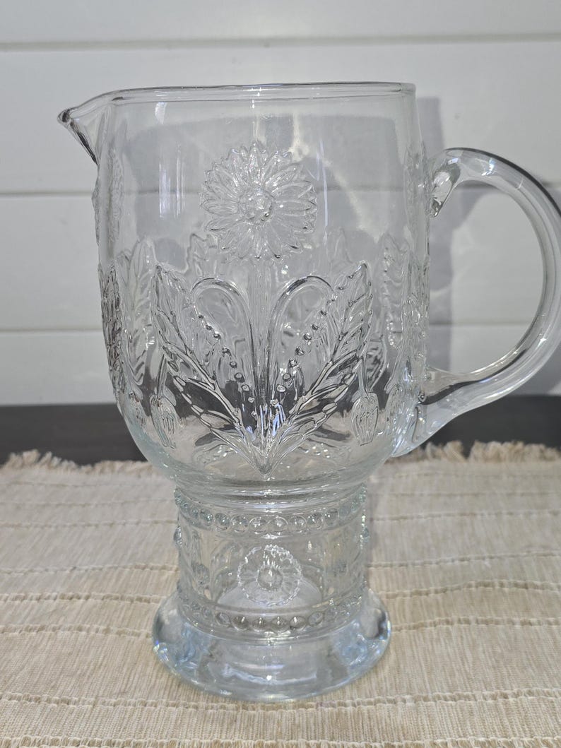 May include: Clear glass pitcher with a floral design. The pitcher features a spout and a handle. The floral design is embossed on the glass, with a large flower in the center and leaves around it. The pitcher sits on a pedestal base.