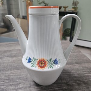 May include: White ceramic teapot with a floral design. The teapot has a ribbed body and a lid with an orange rim. The floral design features blue flowers, green leaves, and a large orange flower.