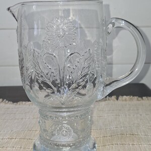 May include: Clear glass pitcher with a floral design. The pitcher features a spout, a handle, and a pedestal base. The floral pattern includes flowers, leaves, and other botanical elements. The pitcher is likely used for serving beverages.