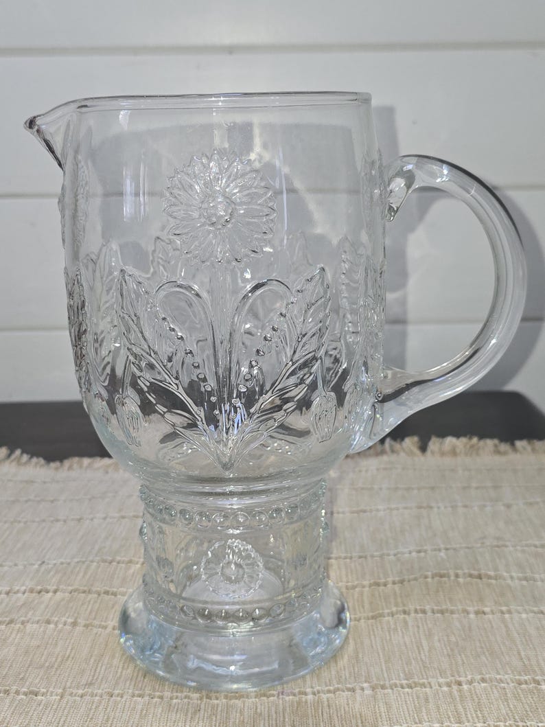 May include: Clear glass pitcher with a floral design. The pitcher features a spout, a handle, and a pedestal base. The floral pattern includes flowers, leaves, and other botanical elements. The pitcher is likely used for serving beverages.