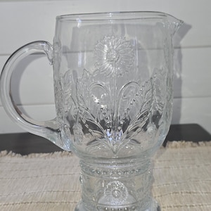 May include: Clear glass pitcher with a handle and a spout. The pitcher features embossed floral designs, including a large flower in the center and smaller flowers and leaves around the sides. The pitcher sits on a pedestal base.
