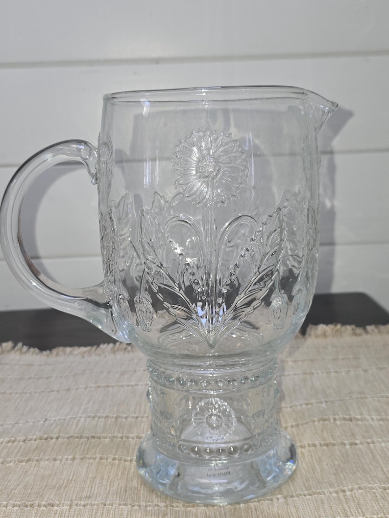 May include: Clear glass pitcher with a handle and a spout. The pitcher features embossed floral designs, including a large flower in the center and smaller flowers and leaves around the sides. The pitcher sits on a pedestal base.