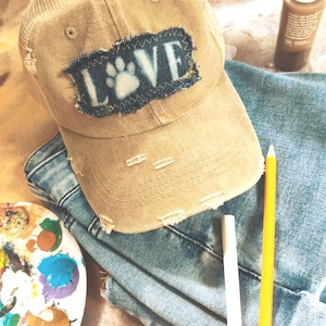May include: Tan baseball cap with a mesh back and a distressed "LOVE" patch featuring a paw print. The cap sits atop a pair of blue jeans, with art supplies like paint and a pencil nearby.