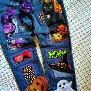 Könnte beinhalten: Dunkelblaue Jeans aus Denim mit verschiedenen Aufnähern und Drucken, darunter eine schwarze Katze, Kürbisse, ein Hexenhut, ein Geist, Totenköpfe und Rosen. Das Wort Magical ist in Schreibschrift gedruckt. Eine lila Wollquaste ist am Bund befestigt.