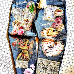 May include: Blue denim jeans featuring a white rope drawstring, lace patches, and various appliqués including cherubs, roses, kittens, and a bird. The text "Dream" and "Angels" is printed on the fabric. The jeans are decorated with intricate lace trim.