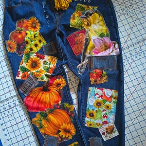 May include: Dark blue denim jeans featuring various fabric patches with sunflowers, pumpkins, apples, and a yellow bird. The word "peace" is written vertically on the left leg, and "Love" is written on the right. A yellow yarn belt is tied at the waist.