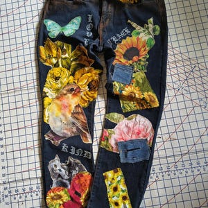 May include: Dark blue denim jeans featuring a yellow braided belt and various fabric patches including a bird, sunflowers, roses, a butterfly, and two cats. The jeans display the words LOVE, PEACE, and KIND in a gothic font style on the fabric.