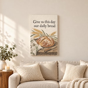 Op de afbeelding: Een rechthoekige wandkunstprint met de tekst Give us this day our daily bread in zwarte letters. De illustratie toont een rond brood op een houten snijplank met tarwearen, een mes en een gestreept stoffen servet.
